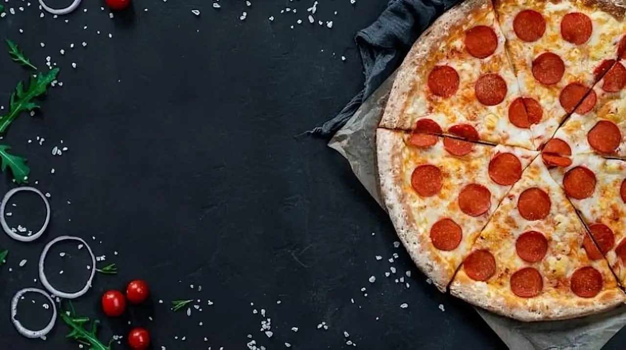 Close-up image of a pepperoni pizza slice on dark background with some green leaves and onion rings scattered
